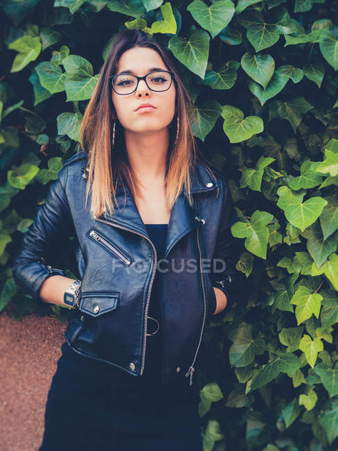 Teen in eyeglasses and leather jacket looking at camera near green foliage of shrub — Stock Photo