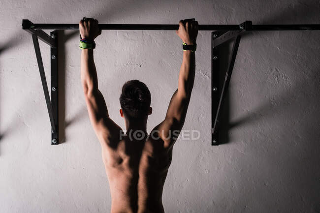 Back view of athletic young shirtless guy hanging on bar near wall in gym — Stock Photo