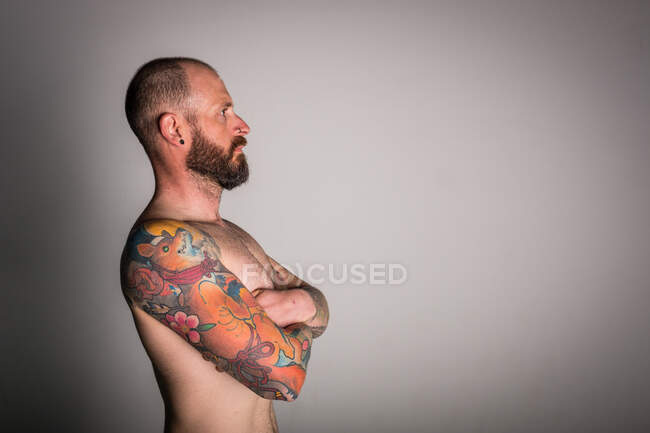 Side view of bearded shirtless hipster with crossed hands and tattoos looking away on grey background — Stock Photo