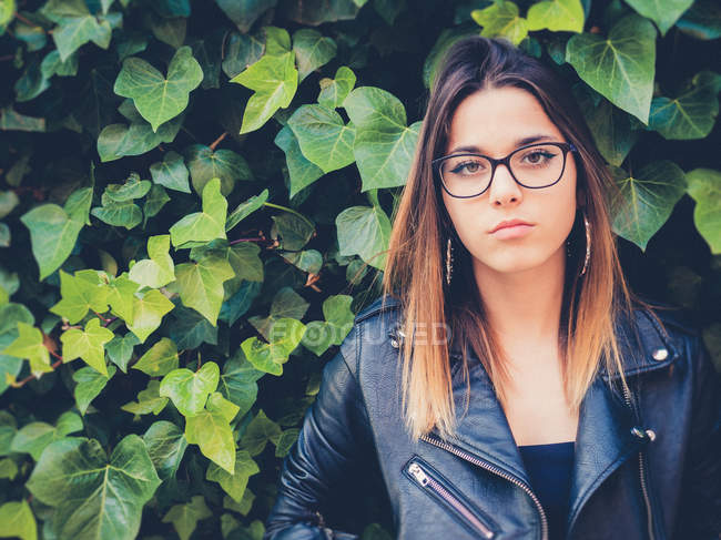 Teen in eyeglasses and leather jacket looking at camera near green foliage of shrub — Stock Photo