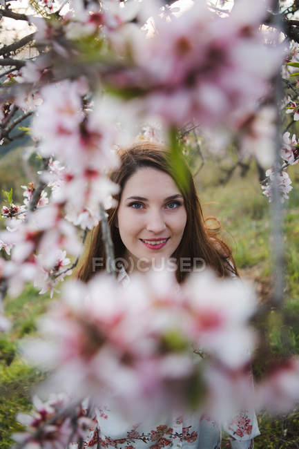 Вид сквозь клумбы цветущего фруктового дерева привлекательной жизнерадостной дамы, глядящей в камеру в саду — стоковое фото