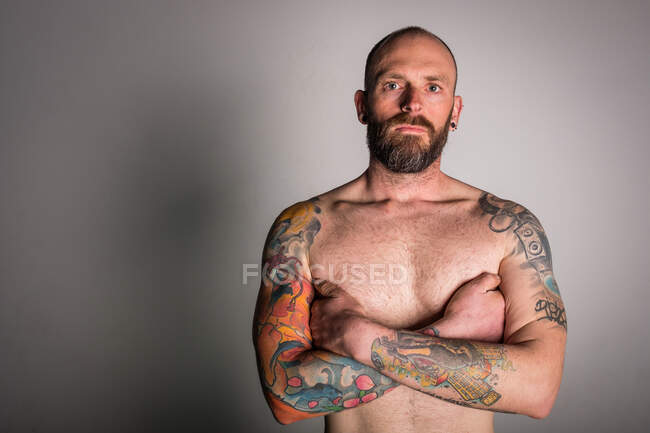 Hipster sin camisa barbuda con las manos cruzadas y tatuajes mirando a la cámara sobre fondo gris - foto de stock