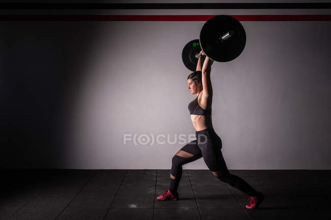 Vista lateral de la joven atlética concentrada dama en ropa deportiva subir la barra por encima de la cabeza en el gimnasio - foto de stock