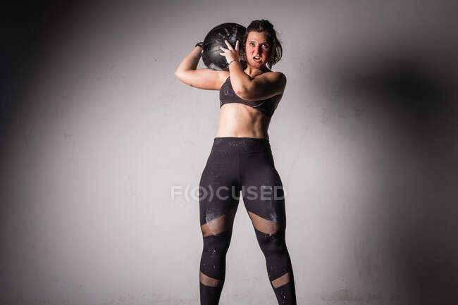 Спортивная молодая сконцентрированная леди в спортивной одежде в меле поднимает мячик тяжелой медицины на плечо в спортзале — стоковое фото