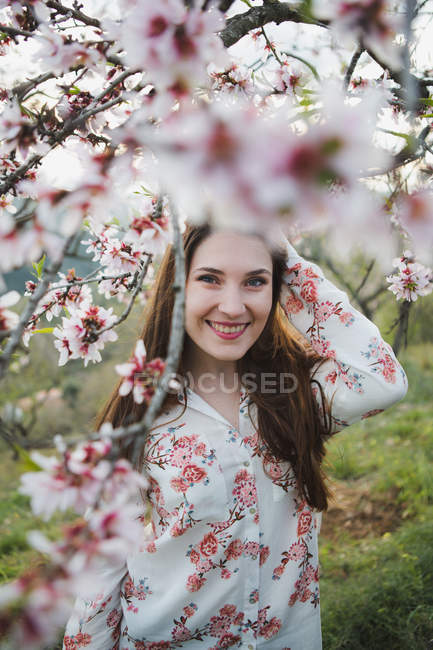 Blick durch Zweige blühender Obstbäume einer attraktiven, fröhlichen Dame, die im Garten in die Kamera blickt — Stockfoto