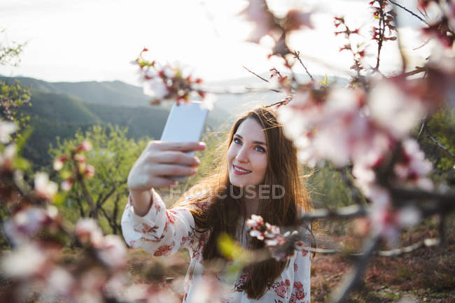 Attrayant gai dame prenant selfie avec téléphone mobile près de fleurs arbre fruitier dans la nature — Photo de stock