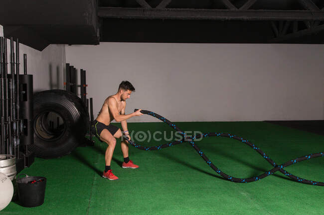Athletic young shirtless guy doing workout with ropes in gym — Stock Photo