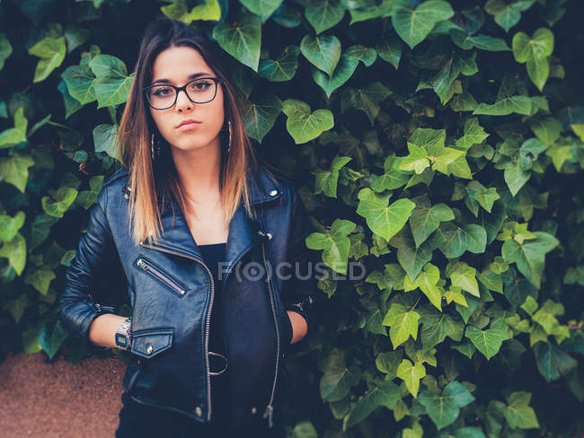 Teenager in Brille und Lederjacke blickt in die Kamera nahe grünem Strauch — Stockfoto