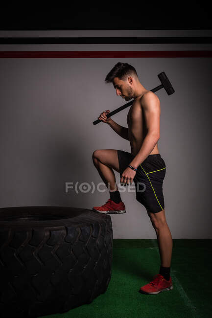 Парень без рубашки, сконцентрированный в спортивной одежде с молотком, бьющим по большой шине в спортзале — стоковое фото
