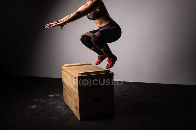 Athletic young lady in sportswear with reached out hands jumping on wooden box in gym on grey background — Stock Photo