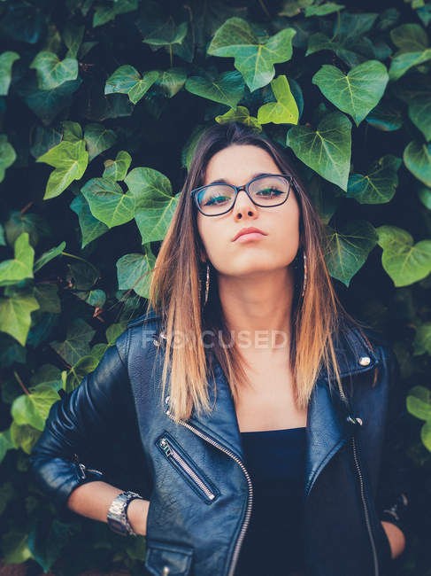 Adolescente en gafas graduadas y chaqueta de cuero mirando a la cámara cerca de follaje verde de arbusto - foto de stock