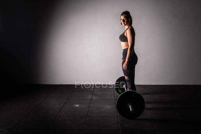 Vue latérale de la jeune femme concentrée athlétique en tenue de sport haussant la barre au-dessus de la tête dans la salle de gym — Photo de stock