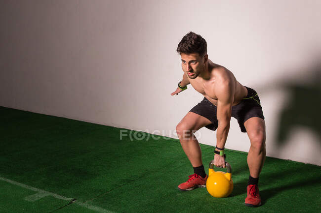 Athletischer junger konzentrierter Kerl in Sportbekleidung, der im Fitnessstudio die Kettlebell hochzieht — Stockfoto