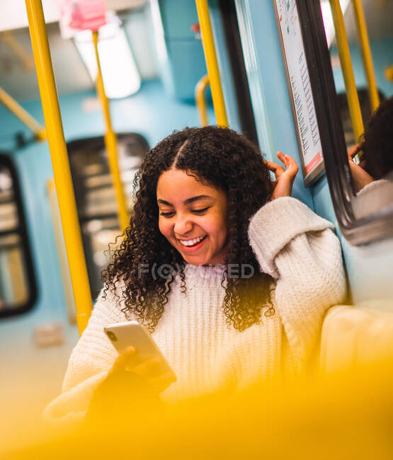 Счастливая привлекательная молодая афроамериканка с кудрявыми волосами просматривает по мобильному телефону в общественном транспорте — стоковое фото