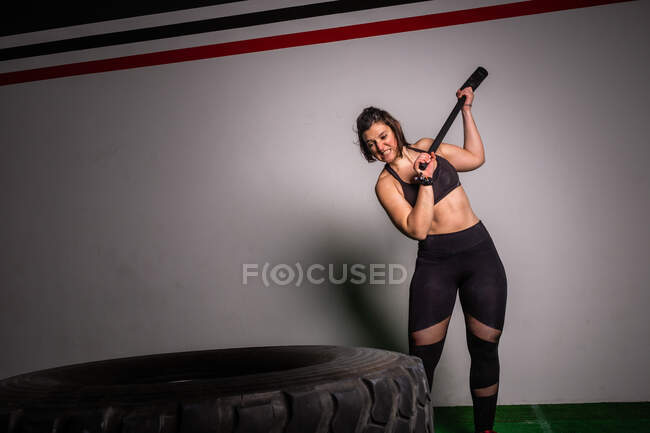 Atlético jovem concentrada senhora no sportswear com martelo batendo no grande pneu no ginásio — Fotografia de Stock