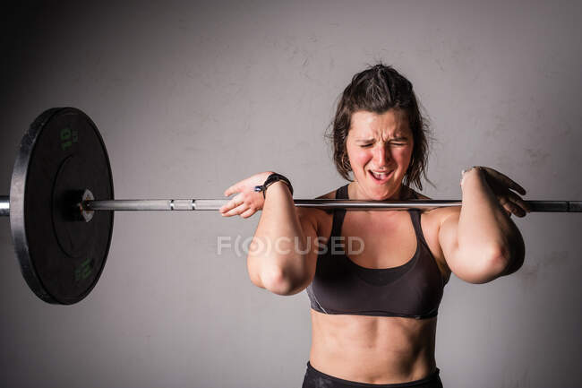 Atlética joven concentrada dama en ropa deportiva subiendo la barra por encima de la cabeza en el gimnasio - foto de stock