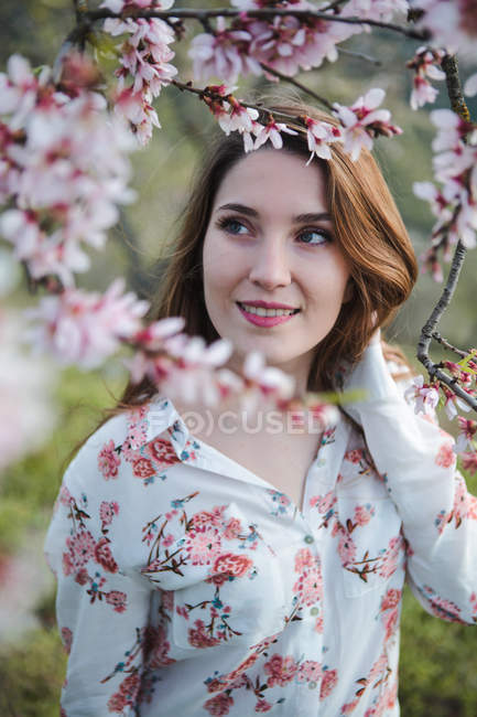 View through twigs of blooming fruit tree of attractive cheerful lady looking away in garden between hills — Stock Photo