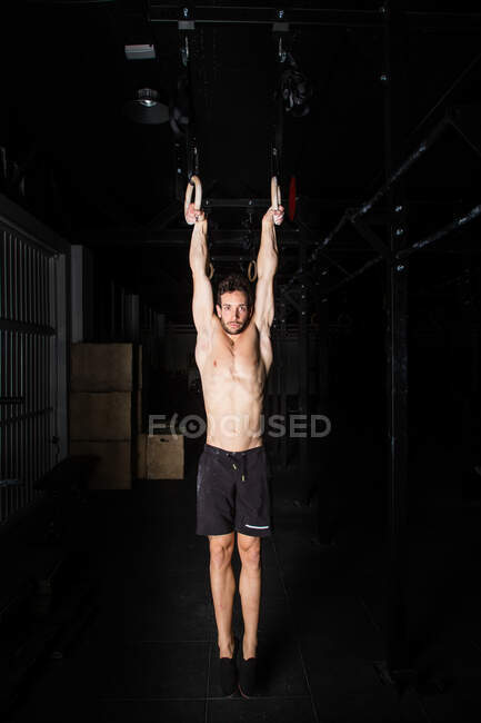 Sportlicher junger Mann ohne Hemd hängt in Turnhalle an Turnringen zwischen Dunkelheit — Stockfoto