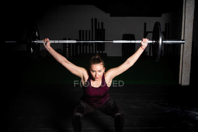 Atletica giovane signora concentrata in abbigliamento sportivo alzando il bilanciere sopra la testa in palestra — Foto stock