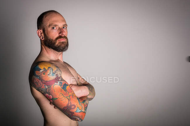 Side view of bearded shirtless hipster with crossed hands and tattoos looking at camera on grey background — Stock Photo