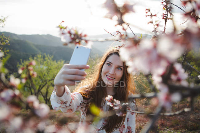 Привлекательная веселая леди делает селфи с мобильным телефоном рядом с цветущим фруктовым деревом в природе — стоковое фото