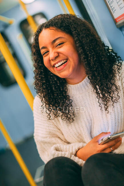 Счастливая привлекательная молодая афроамериканка с кудрявыми волосами просматривает по мобильному телефону в общественном транспорте — стоковое фото