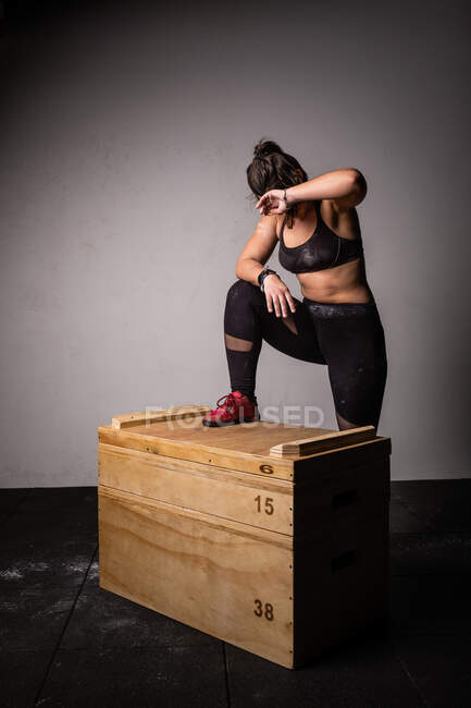 Jeune femme sportive en vêtements de sport avec les mains tendues sautant sur la boîte en bois dans la salle de gym sur fond gris — Photo de stock