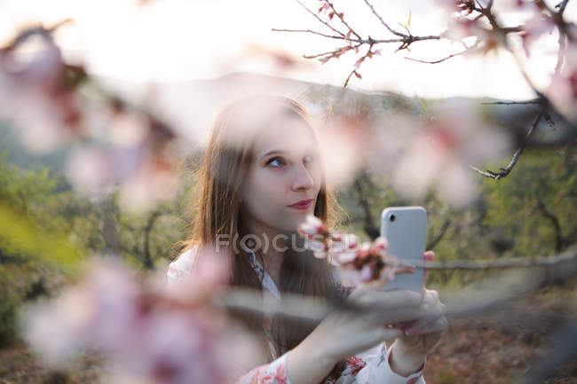 Задумчивая молодая женщина делает селфи с мобильного телефона рядом с цветущим фруктовым деревом в солнечной природе — стоковое фото