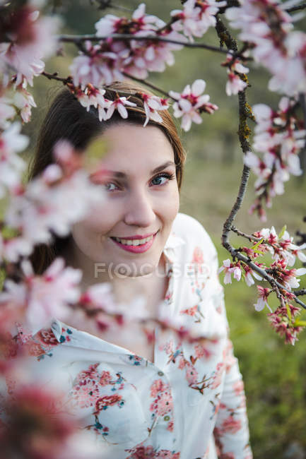 Vista attraverso ramoscelli di albero da frutto in fiore di attraente signora allegra guardando la fotocamera in giardino — Foto stock