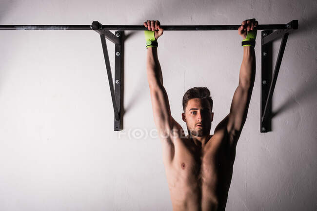 Athletic young shirtless guy hanging on bar near wall in gym — Stock Photo