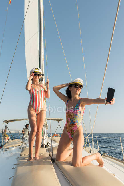 Молоді жінки в сонцезахисних окулярах і капелюхах капітана сідають на мобільний телефон і сидять на палубі дорогого човна, плаваючи на воді в сонячний день. — стокове фото