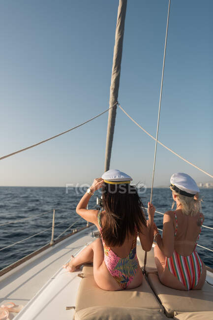 Вид збоку красивих молодих жінок в сонцезахисних окулярах і капелюхах капітана, що сидять на палубі дорогого човна, що плаває на воді в сонячний день — стокове фото