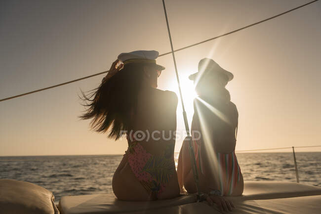 Back view of beautiful young females in sunglasses and captain hats sitting on deck of expensive boat floating on water at sunset — Stock Photo