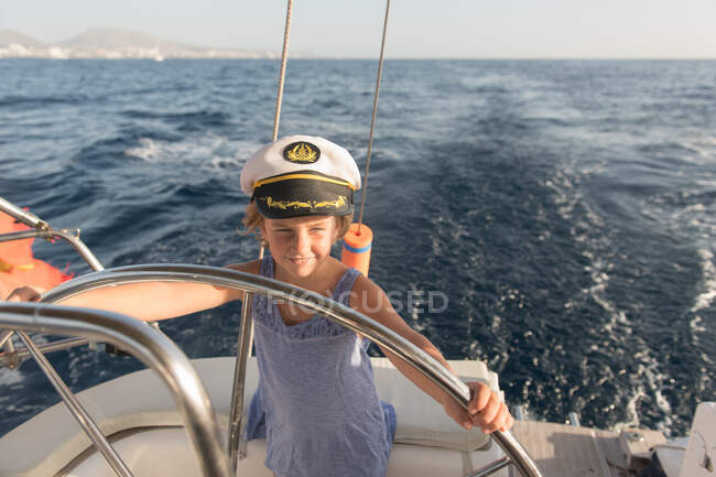 Niño positivo en sombrero de capitán flotando en barco caro en el mar en el día soleado - foto de stock