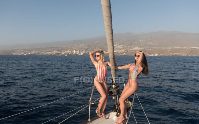 Belles jeunes femmes en lunettes de soleil et chapeaux de capitaine sur le pont latéral d'un bateau coûteux flottant sur l'eau dans la journée ensoleillée — Photo de stock