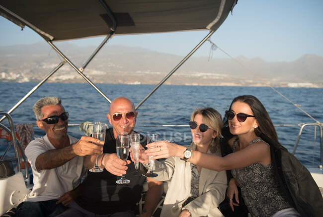 Счастливые мужчины и молодые женщины в солнечных очках, звенящие бокалы шампанского и отдыхающие на дорогой лодке, плавающей на воде в солнечный день — стоковое фото