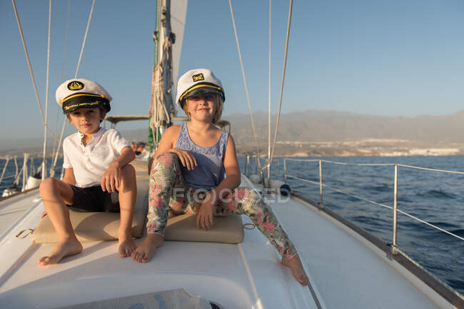 Positive kids in captain hats sitting on deck of expensive boat floating on water in sunny day — Stock Photo