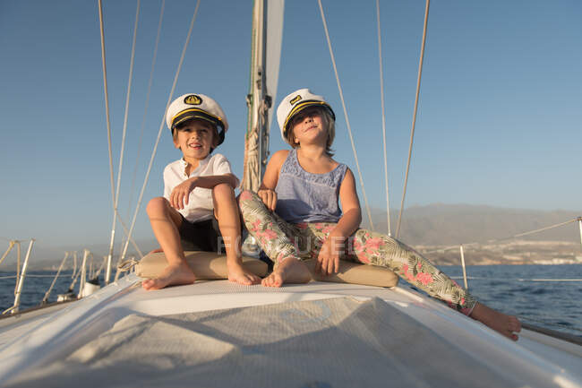 Позитивные дети в капитанских шляпах сидят на палубе дорогой лодки, плавающей по воде в солнечный день — стоковое фото