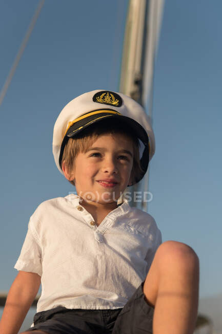 Ragazzo positivo in cappello da capitano seduto sul ponte di barca costosa galleggiante sull'acqua nella giornata di sole — Foto stock