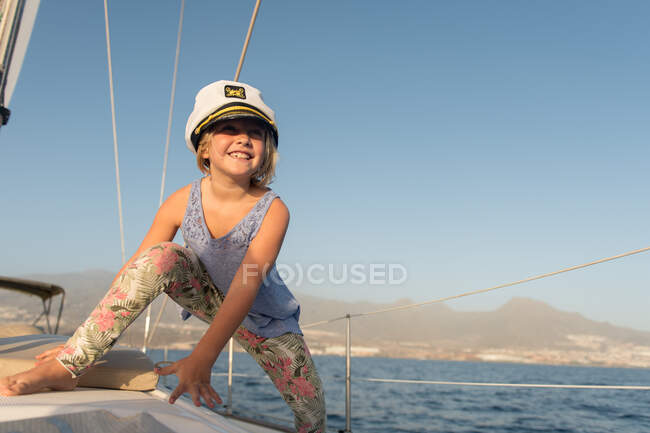 Chica positiva en sombrero de capitán sentado en la cubierta de barco caro flotando en el agua en el día soleado - foto de stock