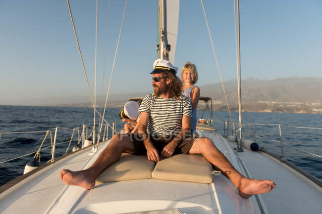 Padre positivo con i bambini felici in cappelli capitano e seduto sul ponte di barca costosa galleggiante sull'acqua nella giornata di sole — Foto stock