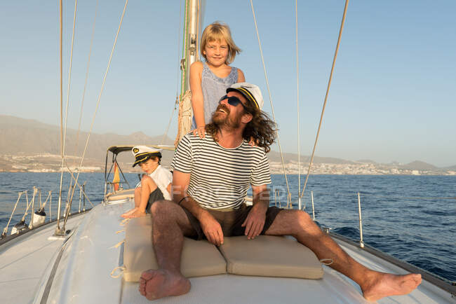 Positive father with happy kids in captain hats and sitting on deck of expensive boat floating on water in sunny day — Stock Photo