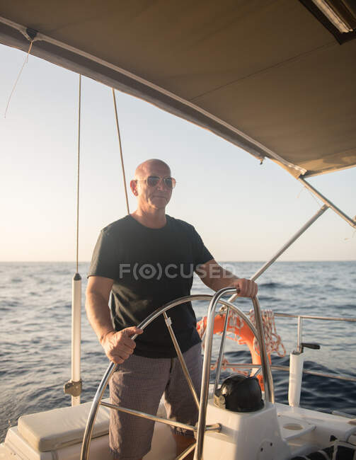 Hombre adulto positivo en gafas de sol flotando en un barco caro en el mar cerca del puerto en un día soleado - foto de stock