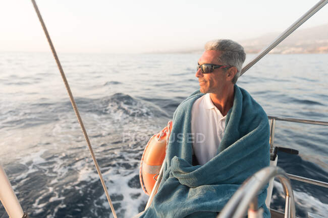 Позитивный взрослый мужчина в солнечных очках и полотенце, сидящий на дорогой лодке, плывущей по морю в солнечный день — стоковое фото