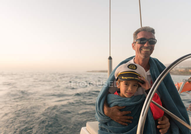 Позитивний батько в сонцезахисних окулярах і рушницях обіймає щасливого хлопця в капелюсі капітана і сидить на дорогому човні, що плаває на воді. — стокове фото