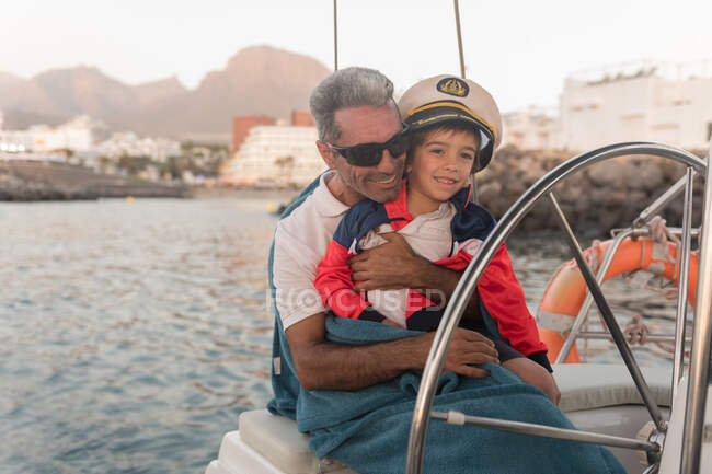 Pai positivo em óculos de sol e toalha abraçando criança feliz em chapéu capitão e sentado em barco caro flutuando na água — Fotografia de Stock