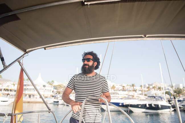 Homme adulte barbu positif dans des lunettes de soleil flottant sur un bateau cher sur la mer près du port par temps ensoleillé — Photo de stock