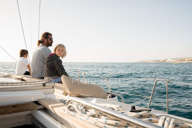 Side view of father with kids sitting on side deck of expensive boat floating on water in sunny day — Stock Photo