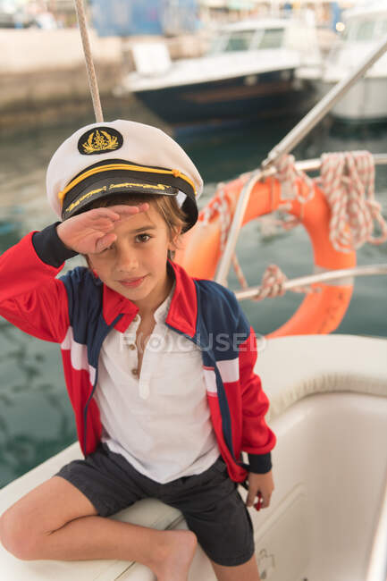 Lustiger Junge mit Kapitänsmütze salutiert und sitzt an Deck eines teuren Bootes, das auf dem Wasser im Dock schwimmt — Stockfoto