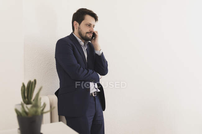 Jeune homme concentré avec la main croisée parlant sur téléphone portable dans la chambre avec plante d'intérieur et réserver à table — Photo de stock